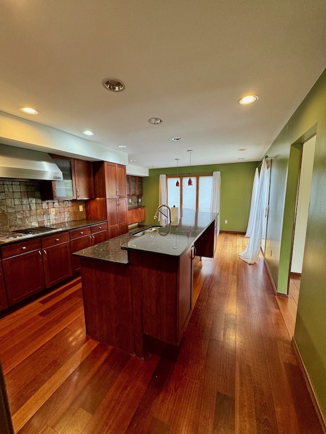 kitchen featuring wall chimney exhaust hood, stainless steel gas cooktop, a kitchen island with sink, sink, and dark hardwood / wood-style floors