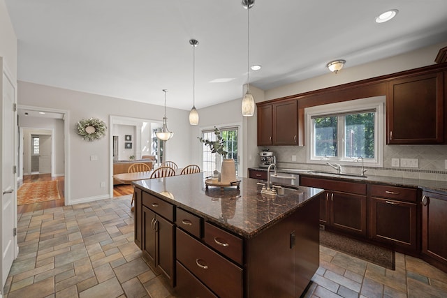 kitchen featuring dark stone counters, sink, an island with sink, tasteful backsplash, and decorative light fixtures