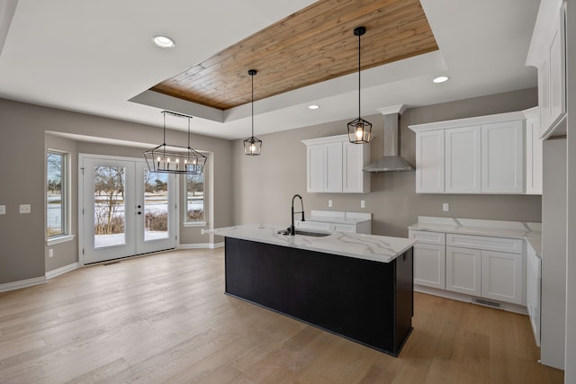 kitchen with sink, a raised ceiling, an island with sink, white cabinets, and wall chimney range hood