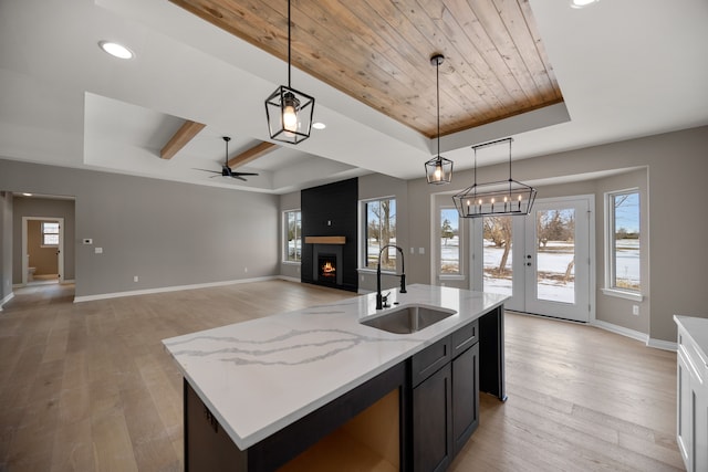 kitchen with sink, light stone counters, a center island with sink, decorative light fixtures, and a raised ceiling