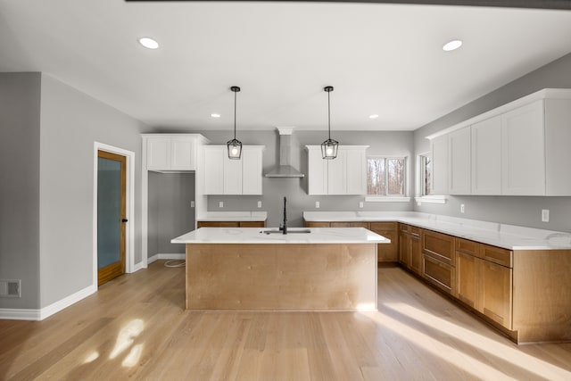 kitchen featuring wall chimney exhaust hood, sink, white cabinetry, decorative light fixtures, and an island with sink