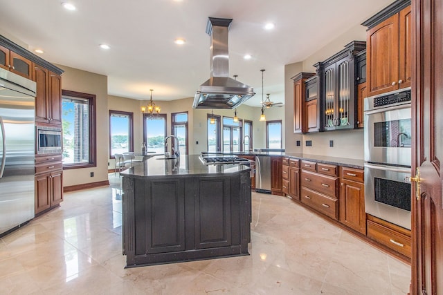 kitchen featuring island exhaust hood, appliances with stainless steel finishes, dark stone counters, a kitchen island with sink, and pendant lighting