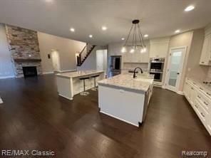 kitchen with double oven, a large fireplace, white cabinets, a center island with sink, and decorative light fixtures