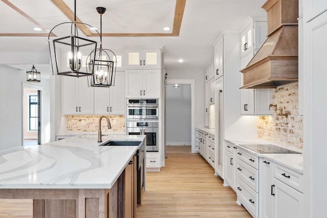 kitchen with light wood-style flooring, glass insert cabinets, stainless steel double oven, premium range hood, and a sink