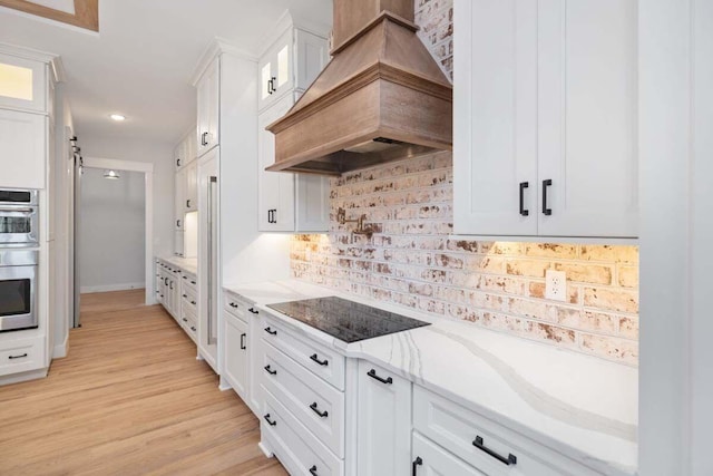 kitchen featuring glass insert cabinets, light stone countertops, black electric stovetop, custom exhaust hood, and white cabinetry