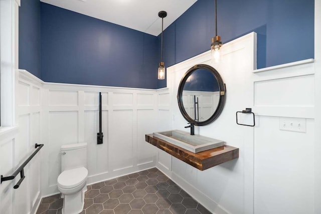 bathroom with wainscoting, toilet, tile patterned flooring, a decorative wall, and a sink