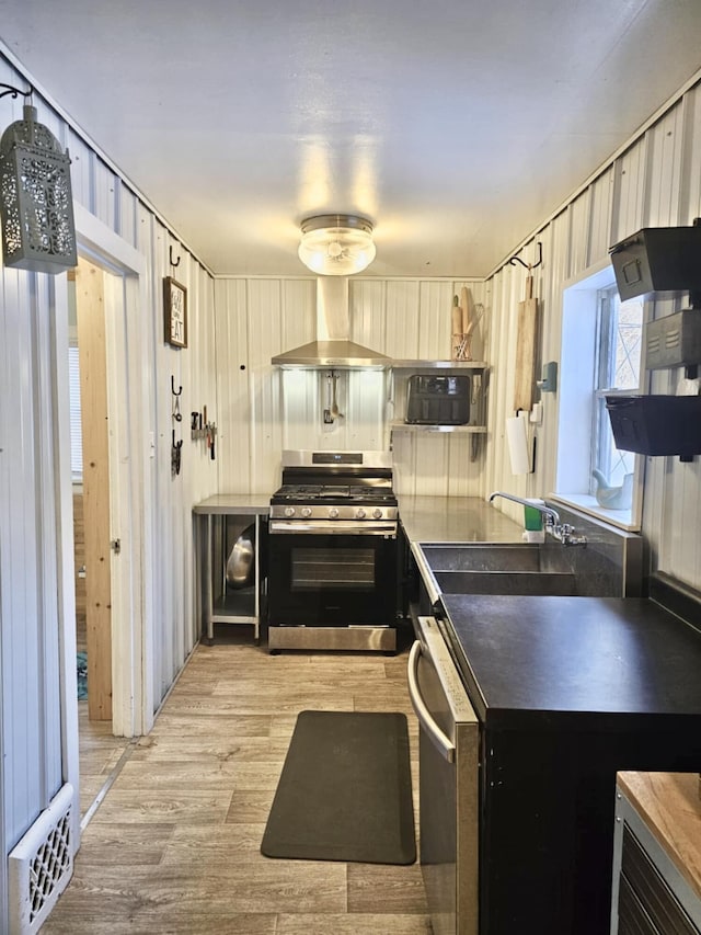 kitchen with dark countertops, appliances with stainless steel finishes, a sink, light wood-type flooring, and wall chimney exhaust hood