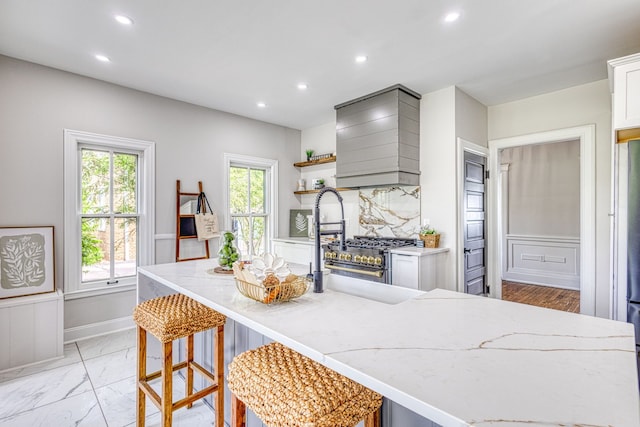 kitchen featuring premium range hood, white cabinets, a center island with sink, decorative backsplash, and a kitchen bar