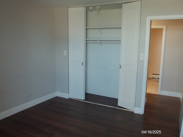 unfurnished bedroom featuring baseboard heating, dark wood-style flooring, a closet, and baseboards