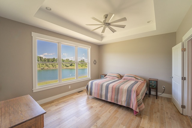 bedroom featuring ceiling fan, light hardwood / wood-style floors, a water view, and a tray ceiling