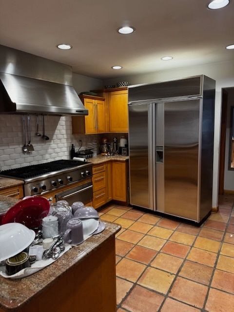 kitchen featuring light tile patterned floors, brown cabinetry, wall chimney exhaust hood, built in refrigerator, and backsplash