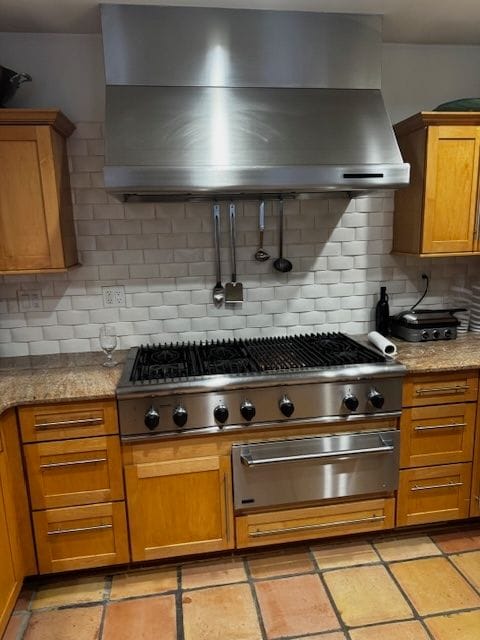 kitchen with tasteful backsplash, brown cabinetry, wall chimney exhaust hood, light stone counters, and stainless steel gas cooktop