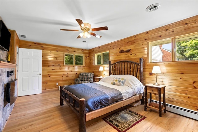 bedroom with ceiling fan, a baseboard radiator, a stone fireplace, wood walls, and light hardwood / wood-style floors