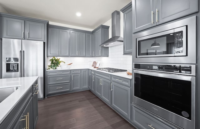 kitchen with dark wood-type flooring, wall chimney exhaust hood, backsplash, gray cabinetry, and appliances with stainless steel finishes