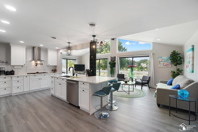 kitchen featuring wall chimney exhaust hood, white cabinetry, dishwasher, pendant lighting, and a kitchen island with sink