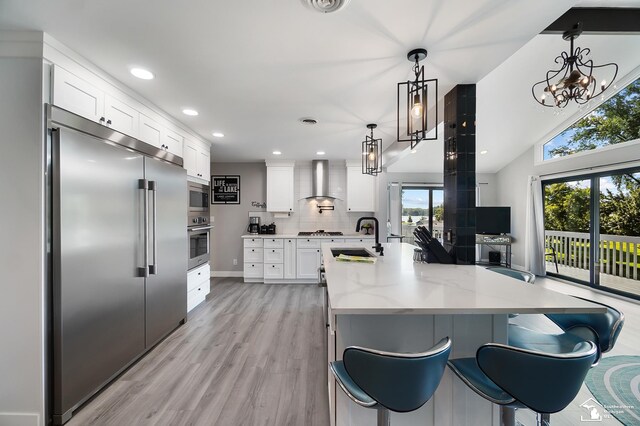 kitchen with wall chimney exhaust hood, sink, built in appliances, hanging light fixtures, and white cabinets