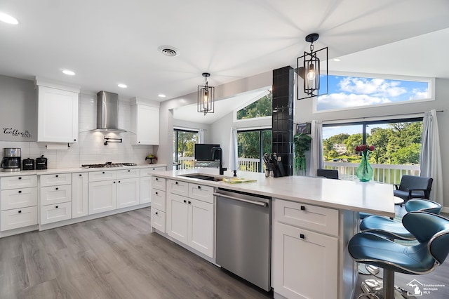 kitchen with hanging light fixtures, white cabinetry, stainless steel dishwasher, and wall chimney range hood