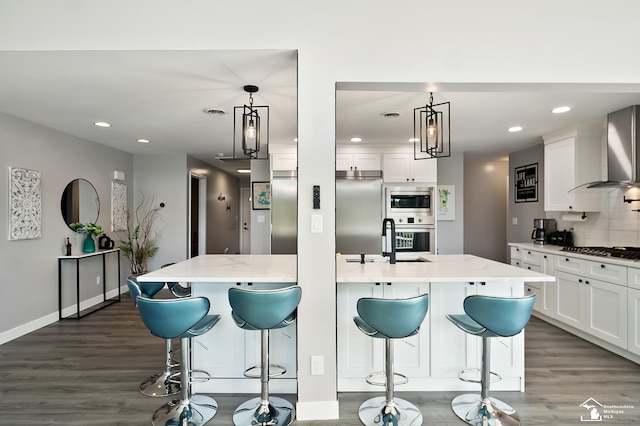 kitchen with decorative light fixtures, a kitchen island with sink, built in appliances, and wall chimney exhaust hood