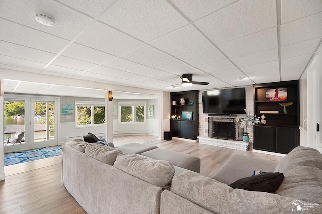 living room with a drop ceiling, a brick fireplace, ceiling fan, and light wood-type flooring