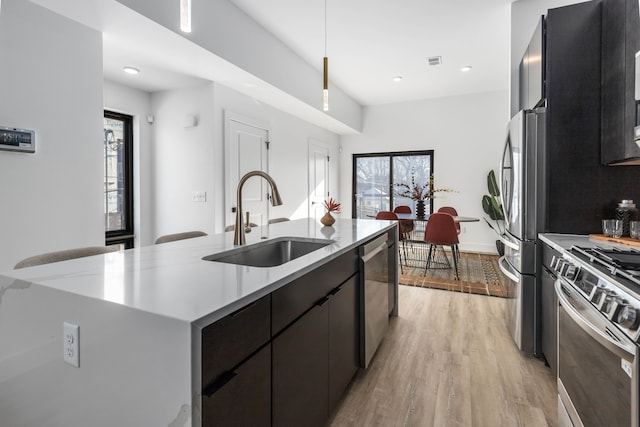 kitchen featuring pendant lighting, a kitchen island with sink, sink, light wood-type flooring, and appliances with stainless steel finishes