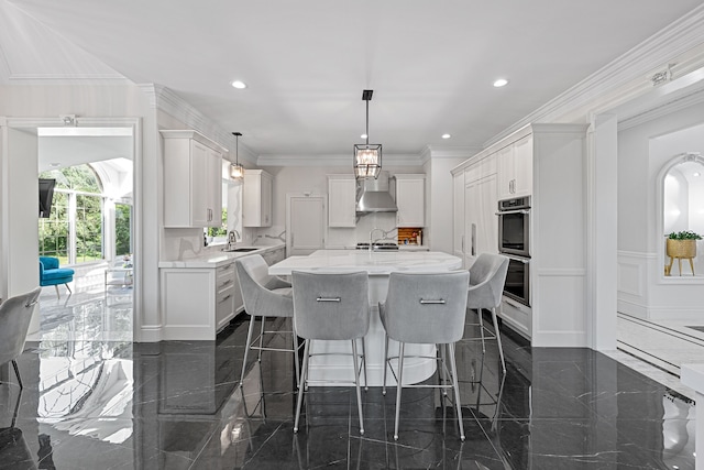 kitchen with wall chimney exhaust hood, stainless steel double oven, a kitchen island with sink, white cabinets, and ornamental molding