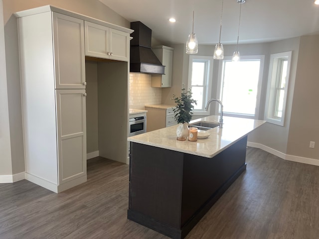 kitchen featuring sink, custom exhaust hood, hanging light fixtures, stainless steel oven, and a center island with sink