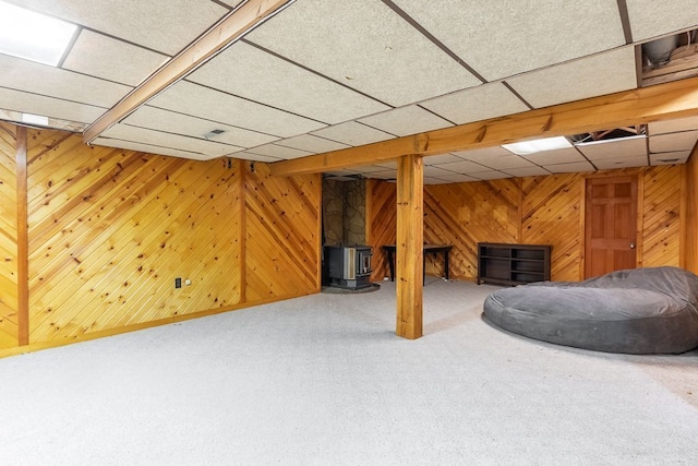 basement featuring a wood stove, wooden walls, carpet, and a paneled ceiling