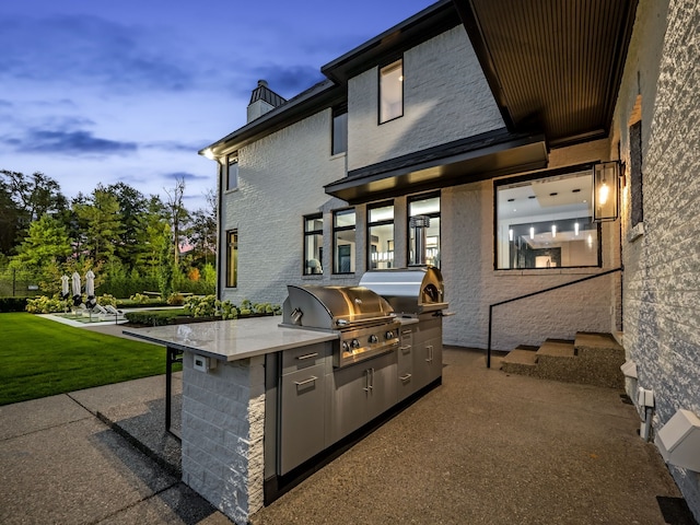 patio terrace at dusk with an outdoor kitchen, an outdoor bar, a lawn, and a grill