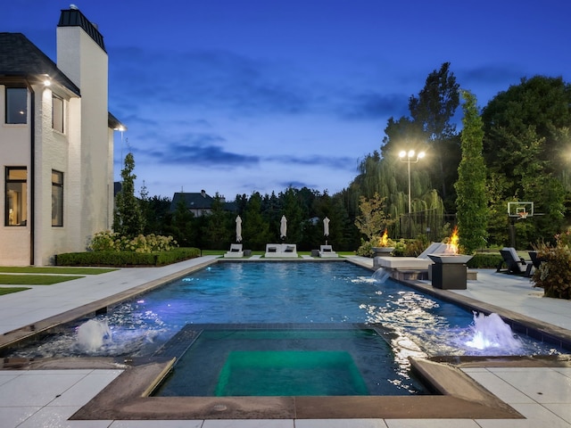 pool at dusk featuring a patio area, an in ground hot tub, pool water feature, and a fire pit