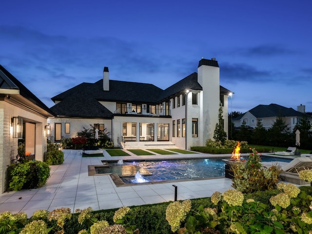 back house at dusk with an outdoor fire pit, pool water feature, a balcony, a swimming pool with hot tub, and a patio