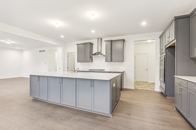 kitchen with an island with sink, sink, wall chimney range hood, and gray cabinetry