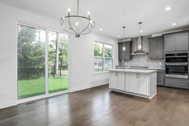 kitchen with gray cabinets, a kitchen island with sink, wall chimney exhaust hood, and hanging light fixtures