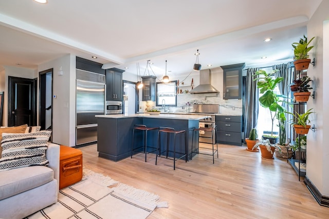 kitchen featuring backsplash, high end appliances, wall chimney range hood, decorative light fixtures, and light hardwood / wood-style floors