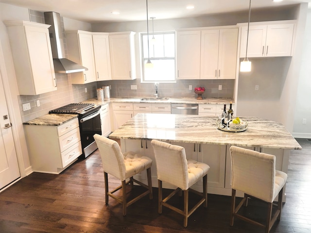 kitchen featuring appliances with stainless steel finishes, wall chimney range hood, white cabinets, a center island, and hanging light fixtures