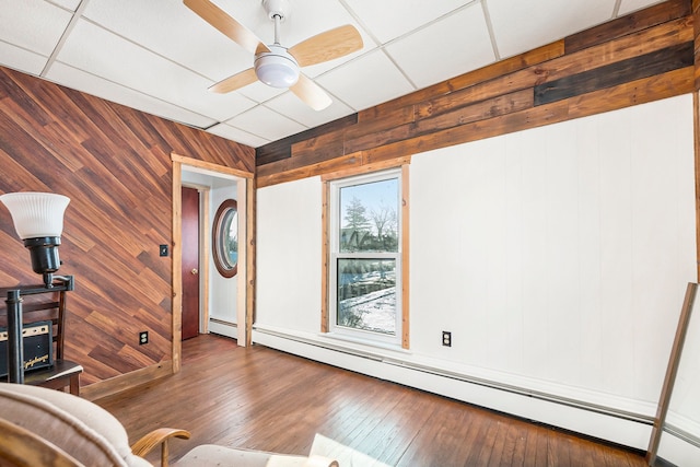 living area with ceiling fan, baseboard heating, a drop ceiling, and wooden walls