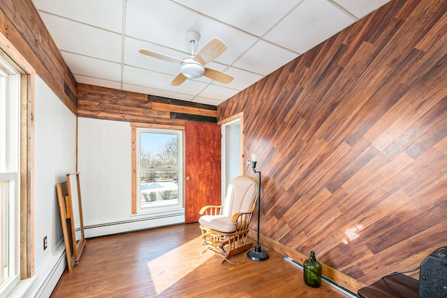 sitting room featuring baseboard heating, dark hardwood / wood-style floors, wood walls, ceiling fan, and a drop ceiling