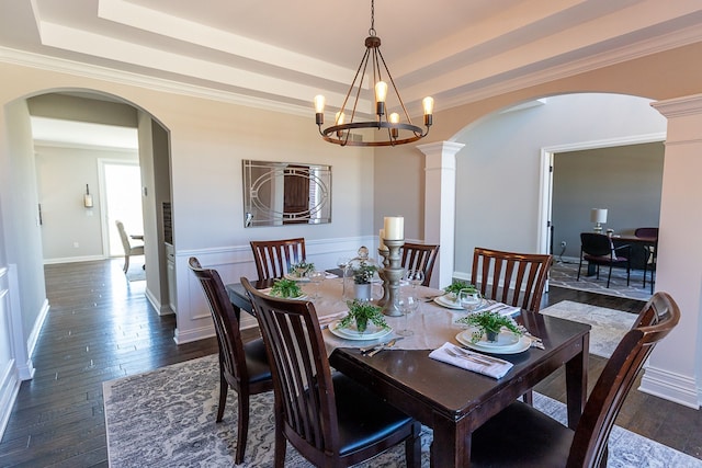 dining area featuring ornate columns, ornamental molding, a tray ceiling, dark wood-type flooring, and an inviting chandelier