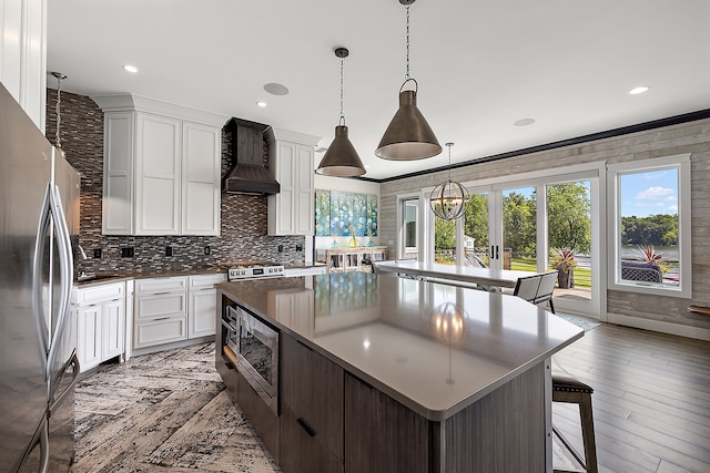 kitchen featuring hanging light fixtures, white cabinetry, appliances with stainless steel finishes, a kitchen island, and custom range hood