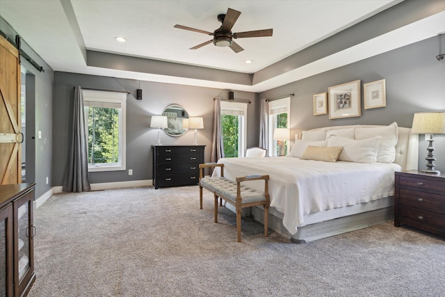 carpeted bedroom featuring ceiling fan, a barn door, a raised ceiling, and multiple windows