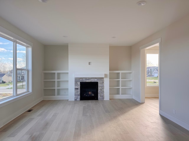 unfurnished living room featuring a wealth of natural light, built in features, light hardwood / wood-style flooring, and a stone fireplace