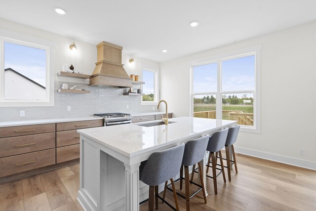 kitchen with a sink, custom range hood, stainless steel gas range, a center island with sink, and light wood-style flooring