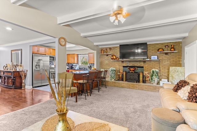 carpeted living room with beamed ceiling, ceiling fan, a wood stove, and ornamental molding