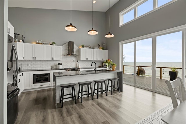 kitchen featuring wall chimney range hood, high vaulted ceiling, fridge, a water view, and white cabinets