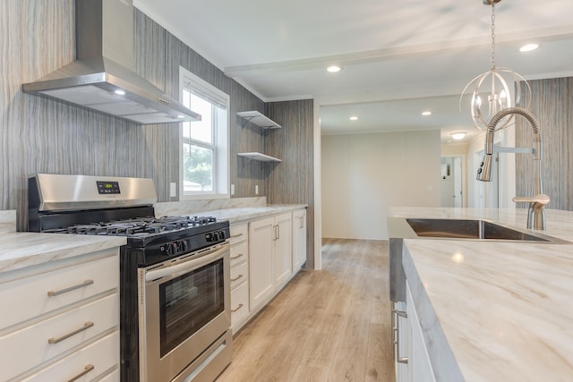 kitchen featuring light wood-type flooring, stainless steel gas range oven, wall chimney range hood, white cabinets, and hanging light fixtures