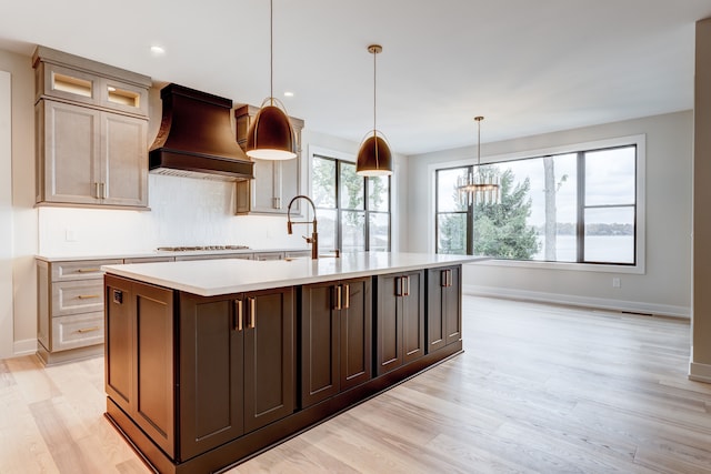 kitchen featuring premium range hood, hanging light fixtures, light wood-type flooring, an island with sink, and gas cooktop