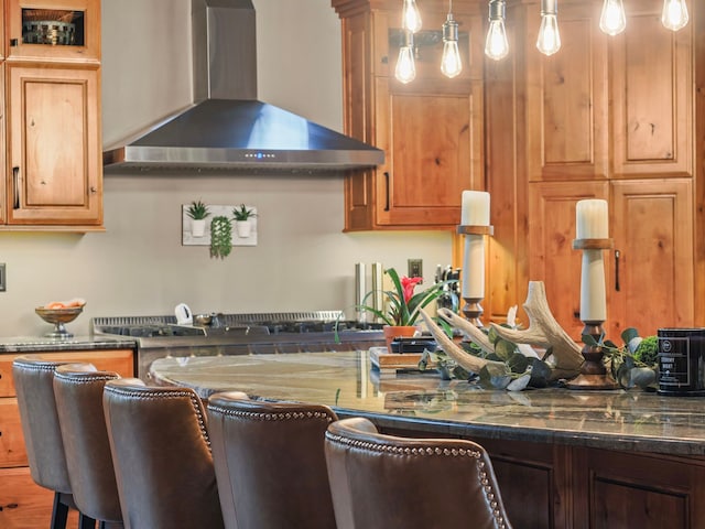 kitchen with hanging light fixtures, wall chimney range hood, a kitchen bar, and dark stone counters