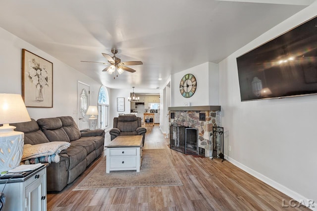 living room with a stone fireplace, ceiling fan with notable chandelier, and light wood-type flooring