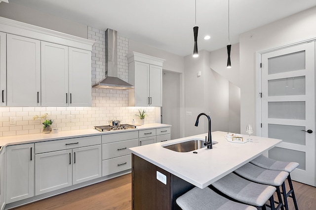 kitchen with white cabinetry, sink, hanging light fixtures, wall chimney range hood, and an island with sink