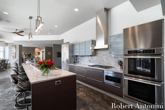 kitchen with wall chimney exhaust hood, backsplash, a breakfast bar area, a kitchen island with sink, and appliances with stainless steel finishes