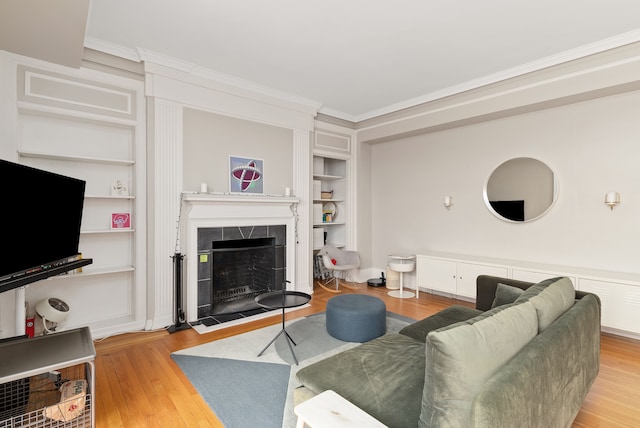 living room featuring a tiled fireplace, crown molding, built in shelves, and wood-type flooring